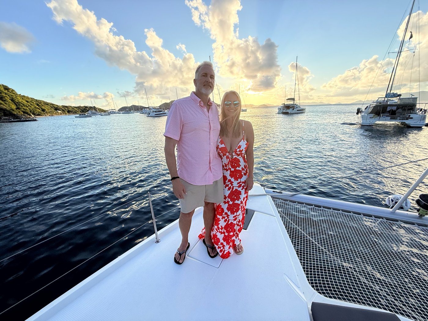 Joe and Danielle Steen smiling together on the foredeck of the catamaran at sunset, a BVI anchorage with other catamarans behind them